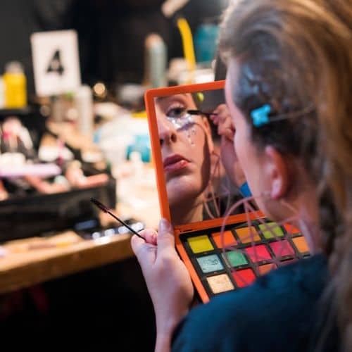 A student from a cosmetology school in Omaha applying makeup in front of a mirror backstage.