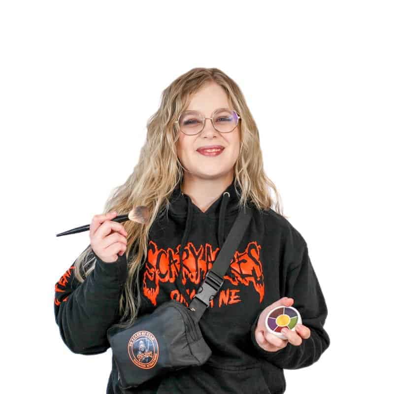 Woman wearing glasses and a black hoodie, holding a makeup brush and a compact, smiling at the camera after her visit to an Omaha cosmetology school.