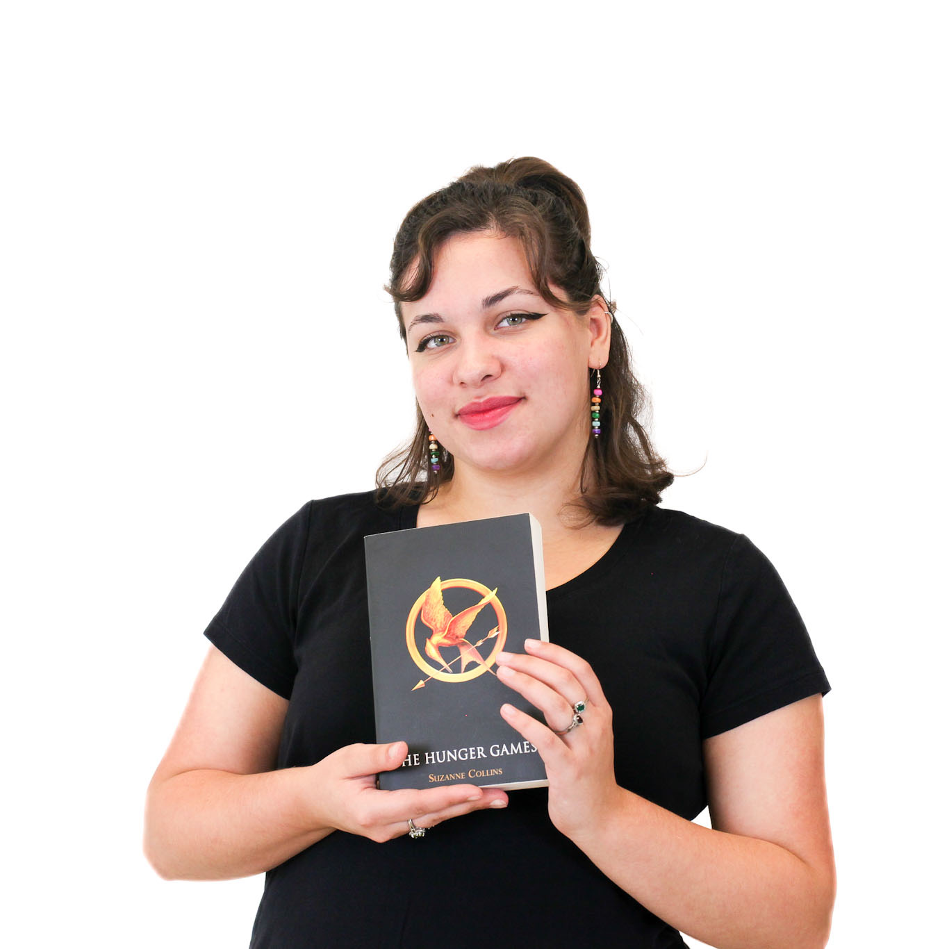 A woman in a black shirt holds a copy of "The Hunger Games" book and smiles at the camera against a plain white background.