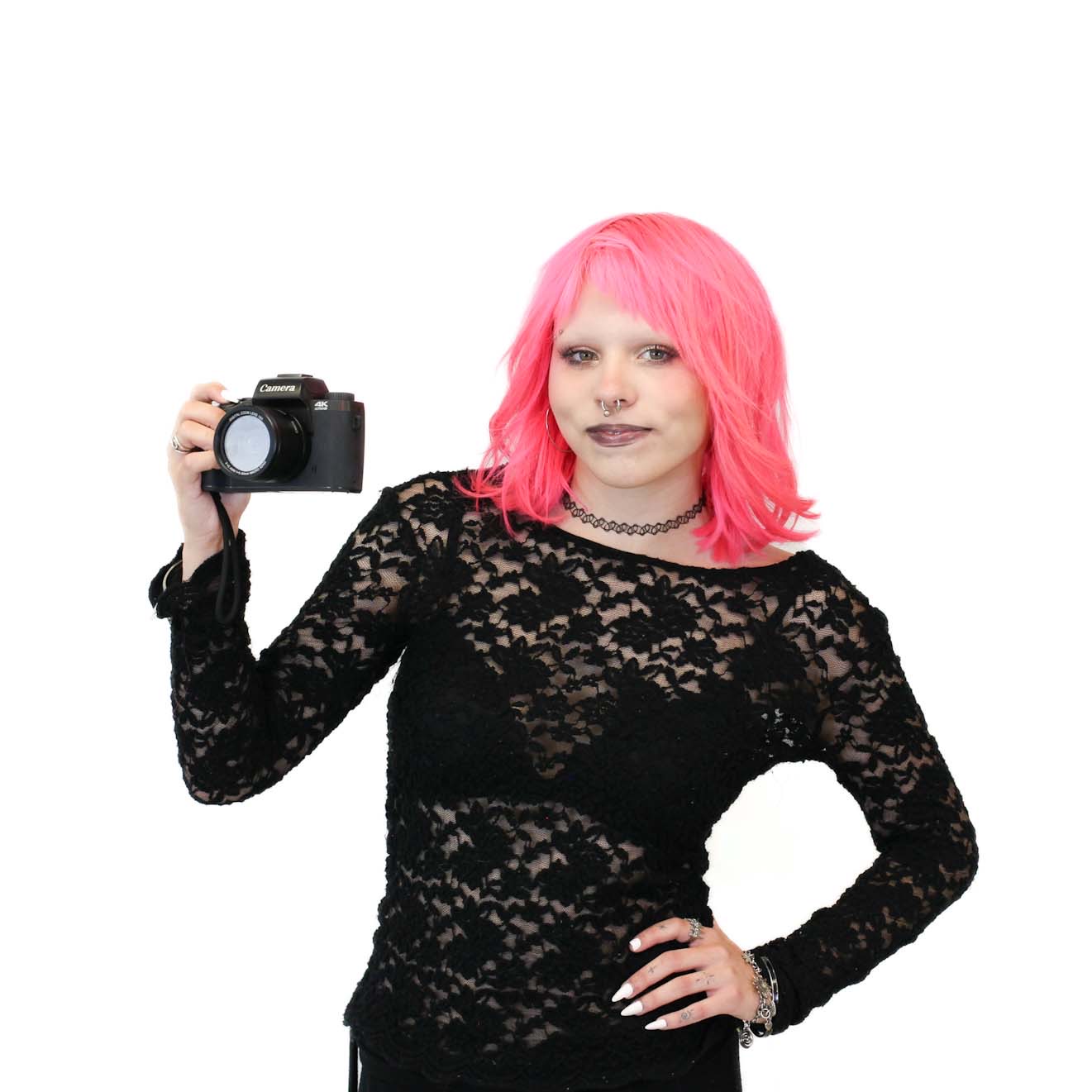 Person with bright pink hair wearing a black lace top holds a camera in their right hand, standing against a plain white background.