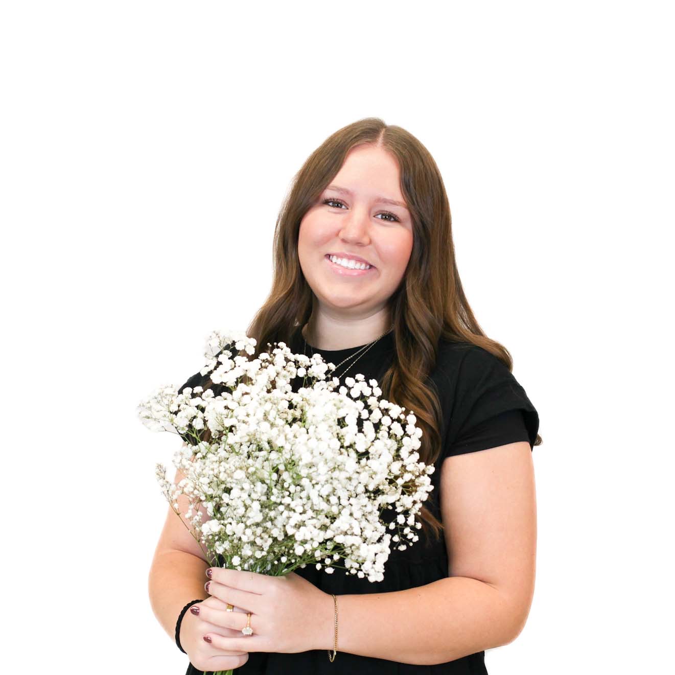 A young woman with long brown hair, wearing a black shirt, smiles while holding a bouquet of white baby's breath flowers against a plain white background.