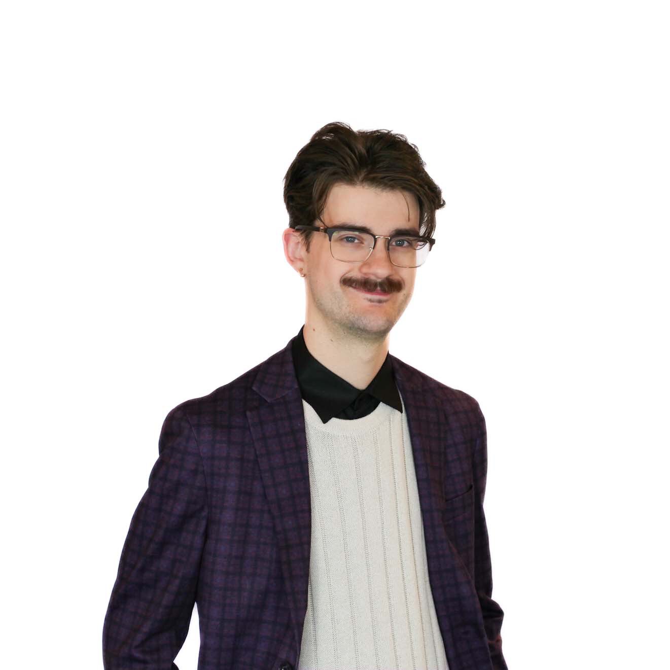 Man with glasses, dark hair, and mustache wearing a purple checked blazer, cream sweater, and black shirt, standing against a plain white background.