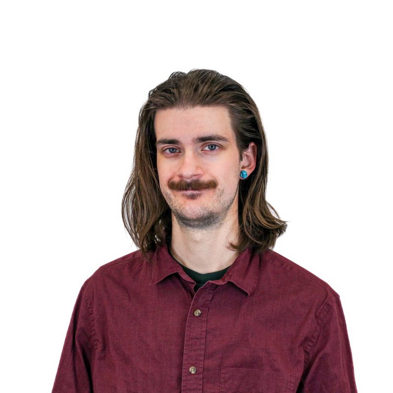 A person with long hair and a mustache, wearing a maroon button-up shirt and blue earring, stands against a plain white background.