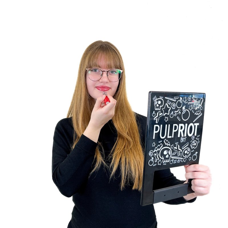 A person with long hair and glasses is applying red lipstick using a mirror from a Pulp Riot kit. They are wearing a black shirt.