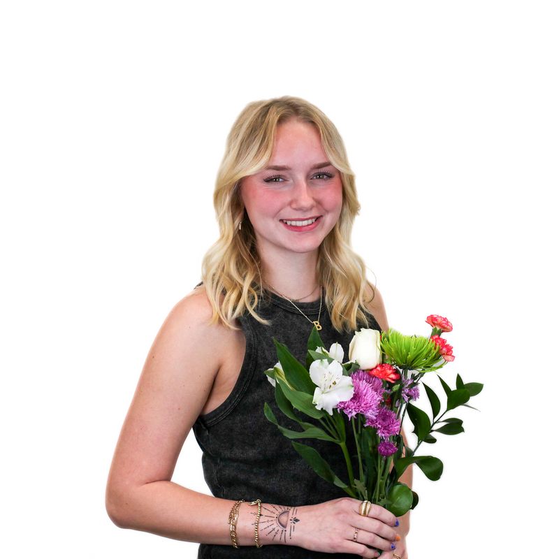 A woman with shoulder-length blonde hair, wearing a sleeveless gray top, holds a bouquet of colorful flowers against a white background.