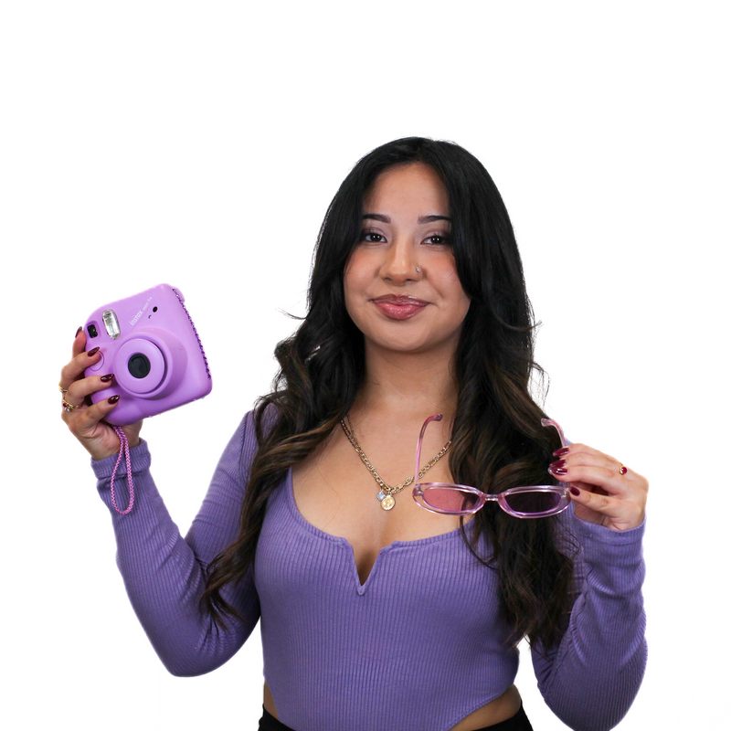 A woman with long dark hair holds a purple instant camera in one hand and a pair of glasses in the other. She is wearing a long-sleeved purple top.