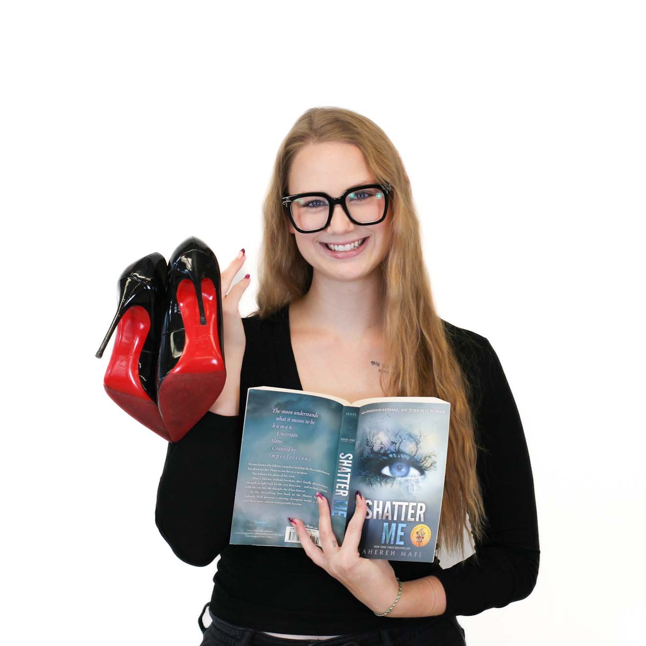 A woman with long blonde hair and glasses holds black high-heeled shoes in one hand and an open copy of the book "Shatter Me" in the other, standing against a white background.