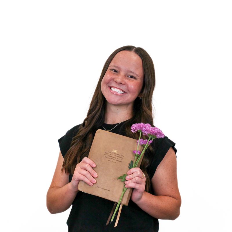 A woman with long brown hair smiles, holding a tan book in one hand and purple flowers in the other, standing against a plain white background.