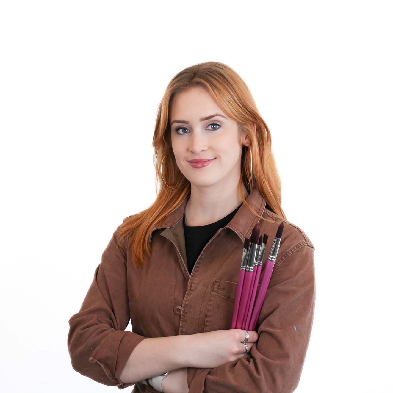 A woman with long red hair stands against a white background, wearing a brown jacket and holding four paintbrushes in her arm.