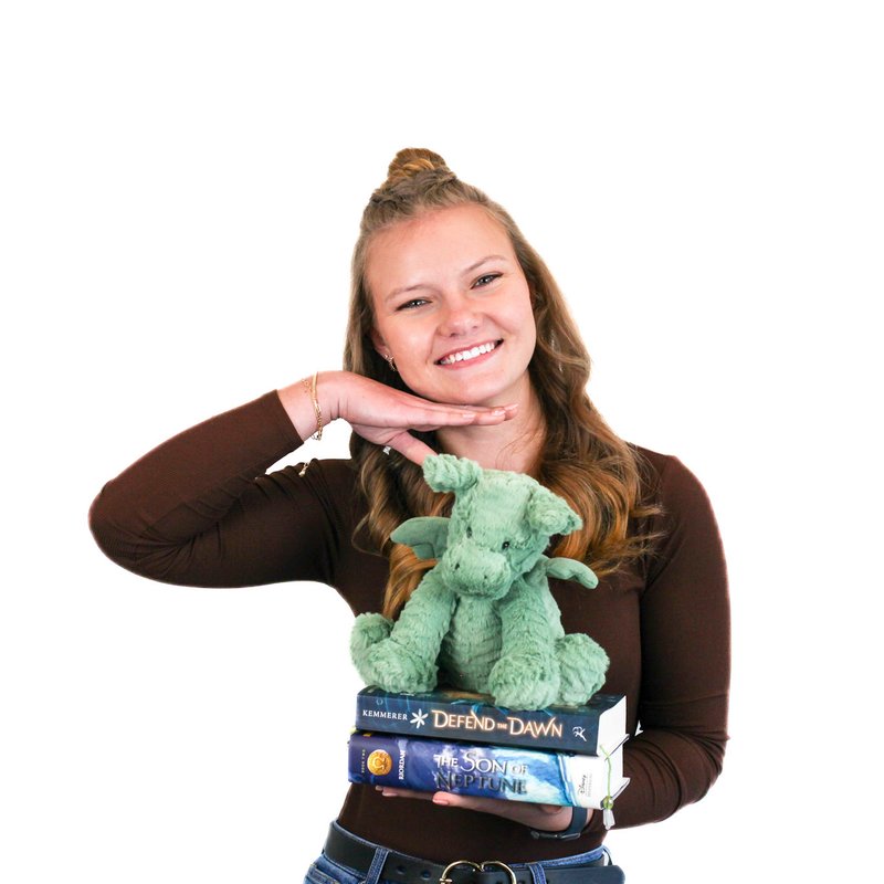 Smiling person with a topknot holds a green plush dragon and two books titled "Defend the Dawn" and "The Son of Neptune," against a white background.