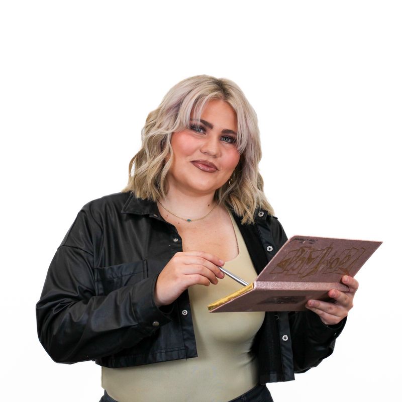 A person with blonde hair holds a makeup palette in one hand and a brush in the other, smiling. They are wearing a black jacket over a light-colored top, standing against a plain white background.