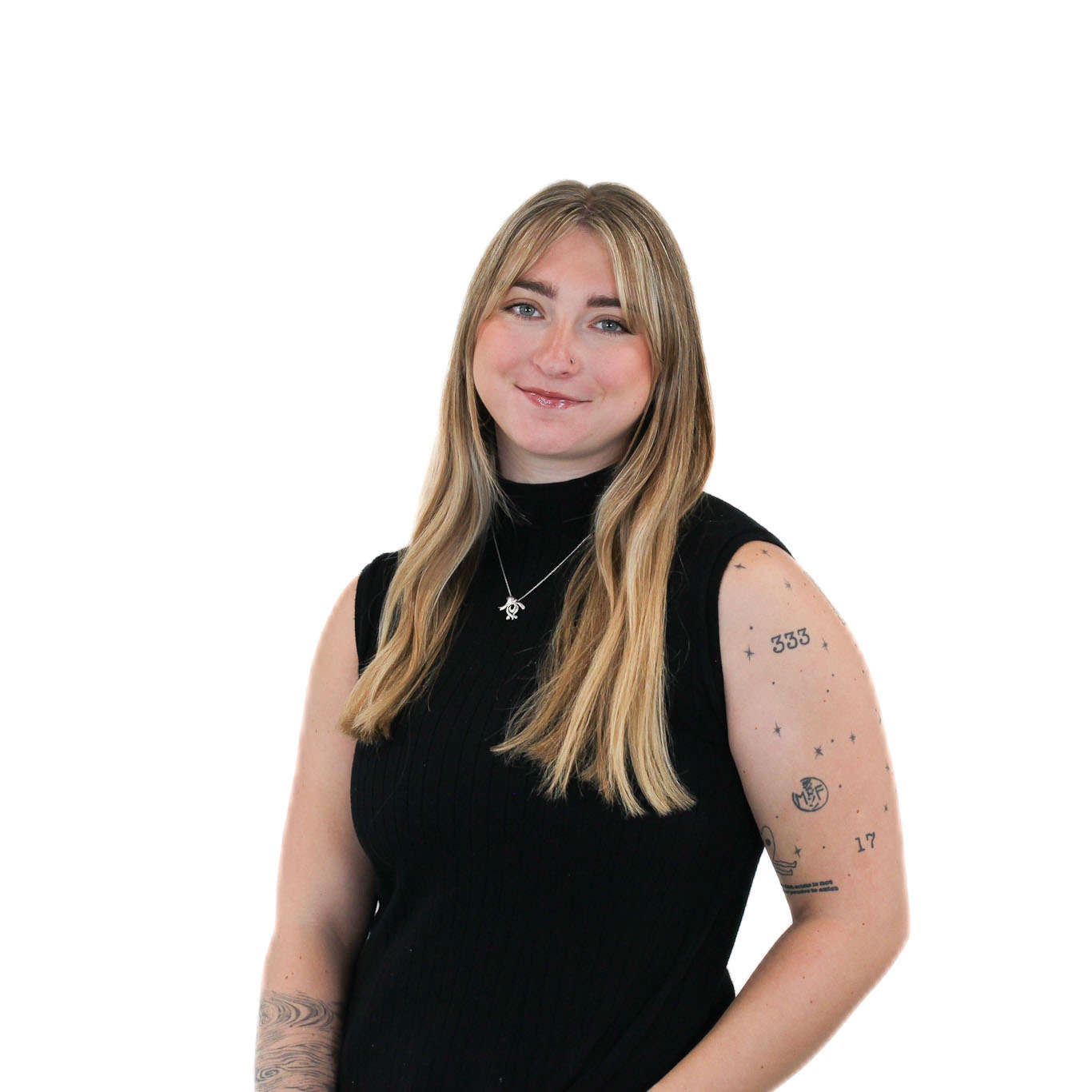 A woman with long blonde hair wearing a black sleeveless top stands against a white background. She has visible tattoos on her left arm and is wearing a necklace.