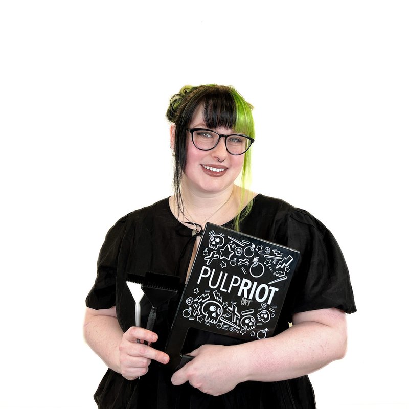 A person with green and black hair holds a Pulp Riot box and a brush, smiling, against a white background.