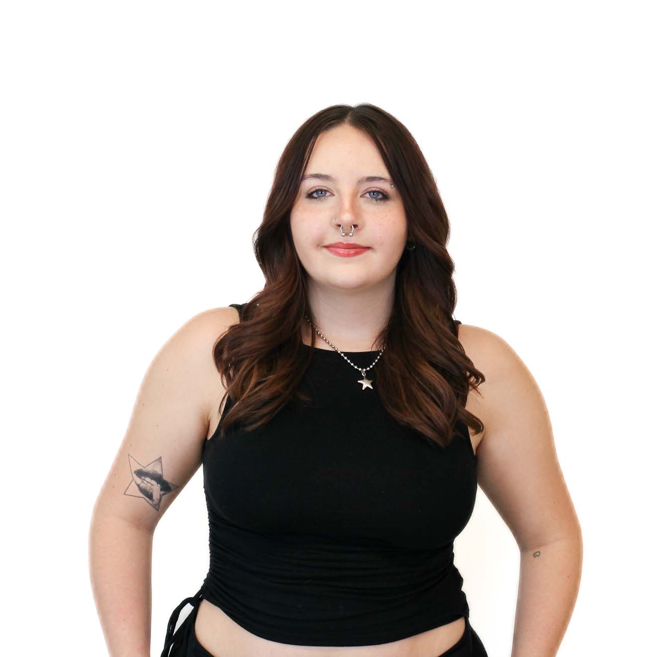 Woman with long brown hair, wearing a black sleeveless top, silver necklace, and septum piercing, standing against a plain white background. She has a star tattoo on her upper arm.