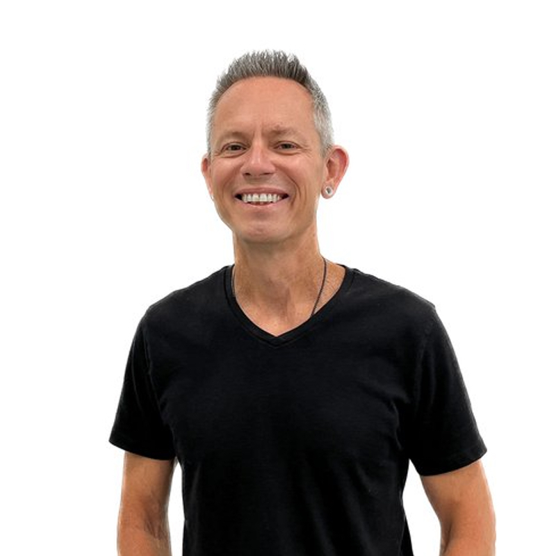 Person with short, gray hair and pierced ears smiling, wearing a black V-neck T-shirt, against a white background.