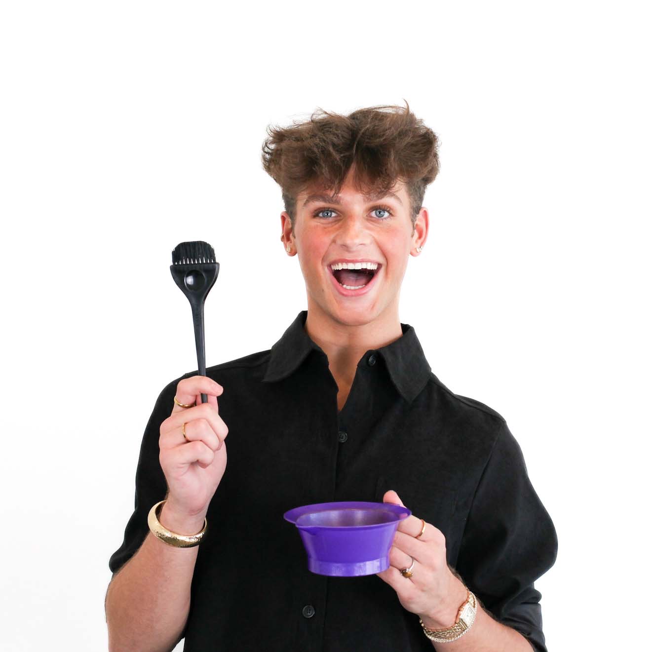 Smiling person in a black shirt holds a hair dye brush in one hand and a purple mixing bowl in the other, standing against a plain white background.