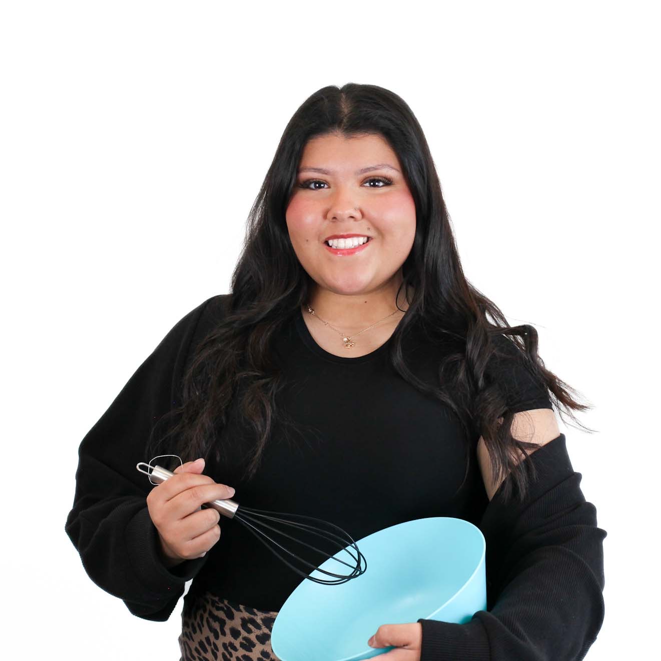 A person with long dark hair, wearing a black top, holds a whisk and a light blue mixing bowl, standing against a plain white background.
