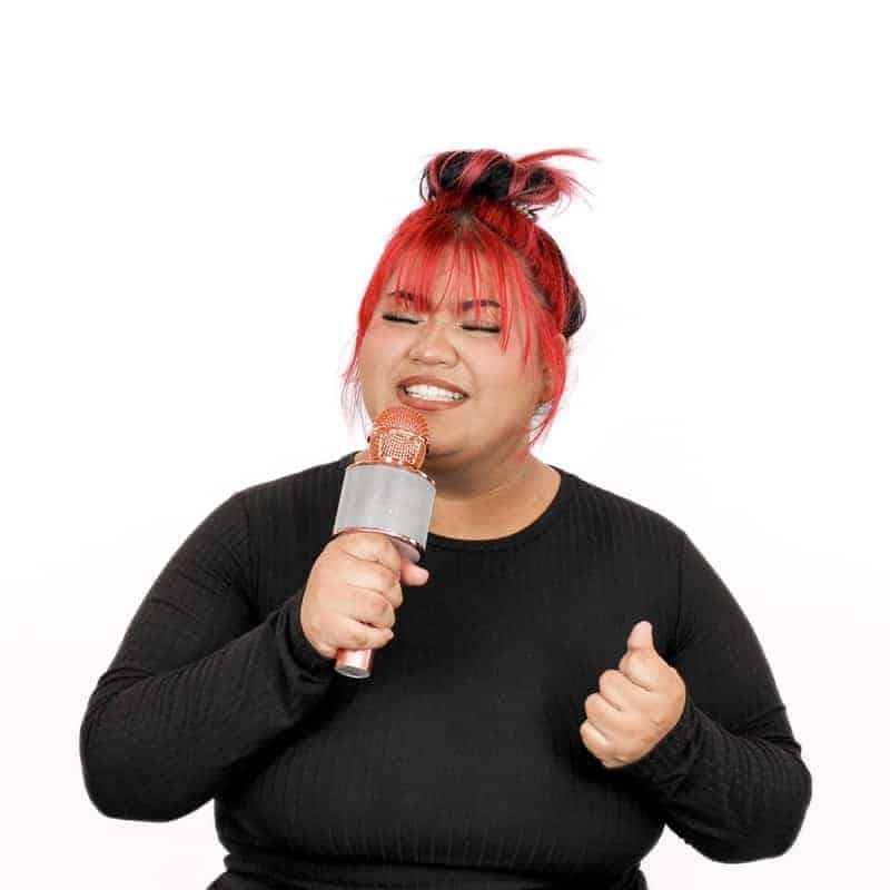A person with red hair, styled by a top cosmetology school, singing into a microphone with a joyful expression.