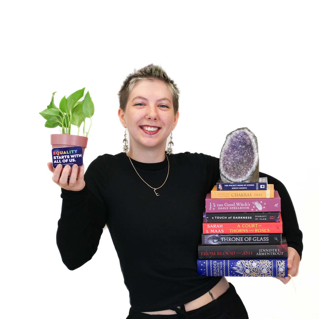 A smiling person holds a small potted plant in one hand and a stack of books topped with a purple geode in the other, against a white background.