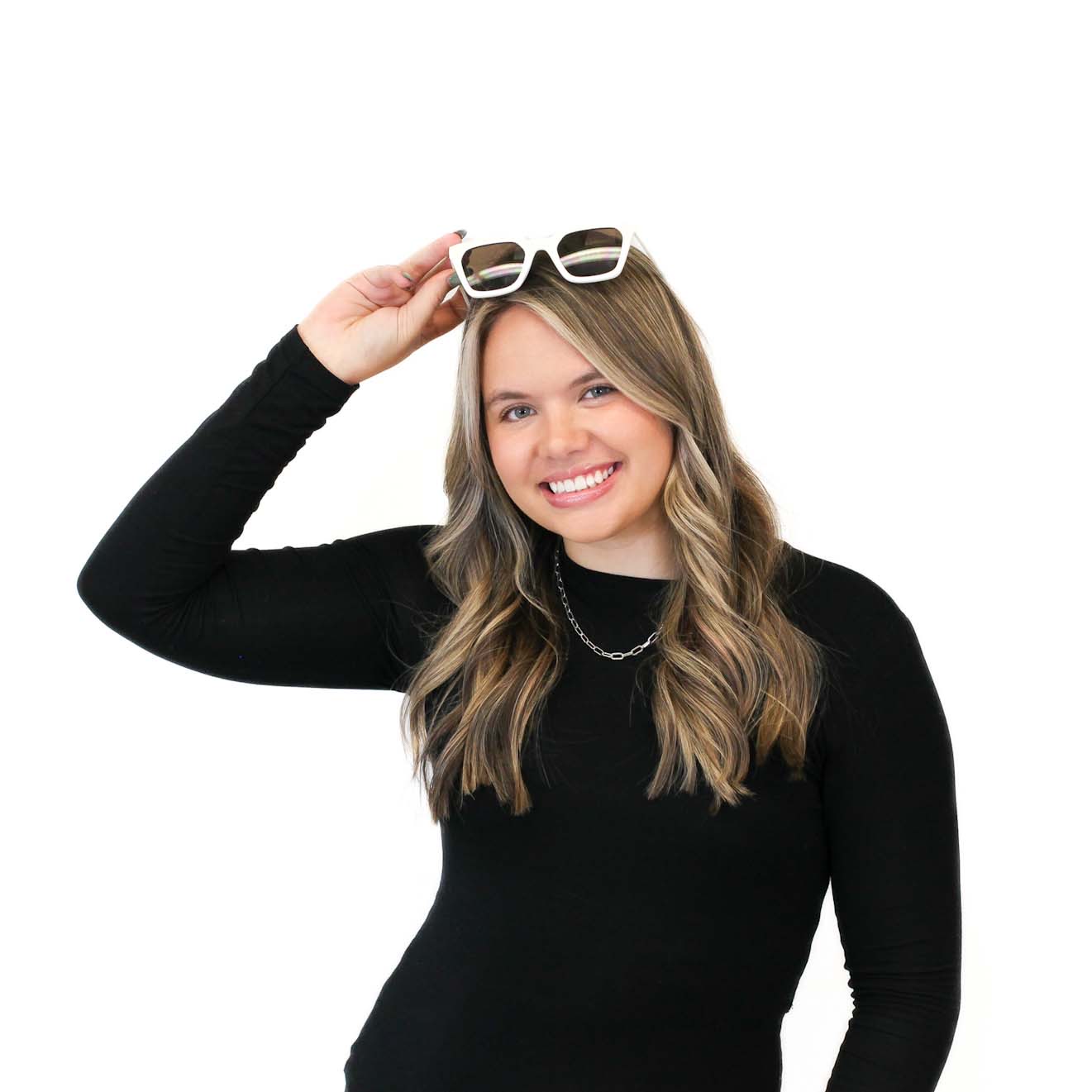 Woman with long blonde hair wearing a black top smiles while holding white sunglasses above her head against a white background.