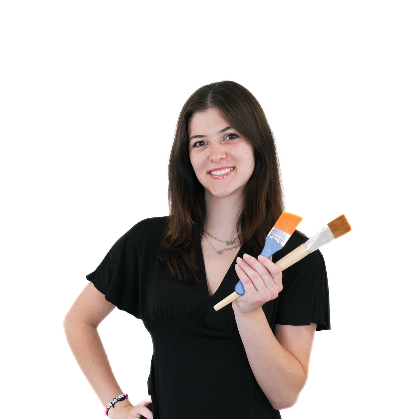 A woman in a black shirt stands against a white background, smiling and holding two paintbrushes in her right hand.
