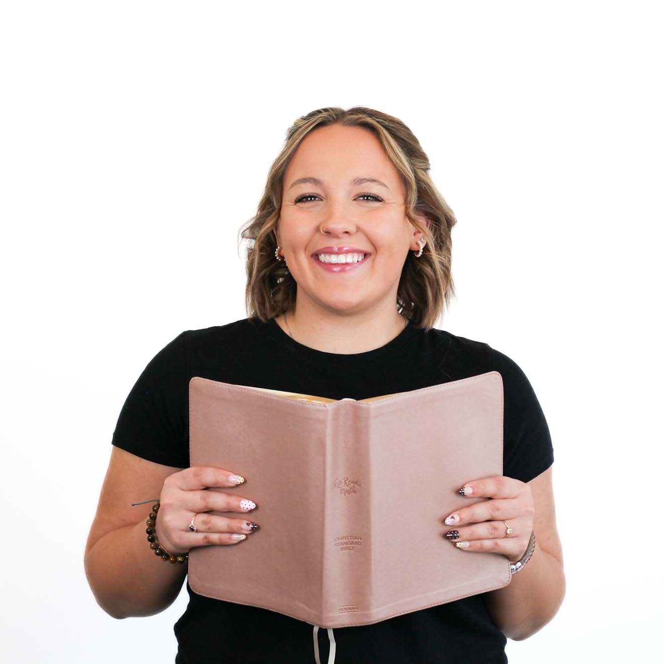 A person smiling and holding an open book with a light pink cover, wearing a black shirt, standing against a white background.