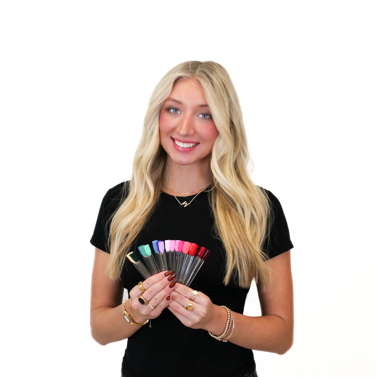 A woman with long blonde hair in a black shirt holds a fan of colorful nail polish samples against a white background.