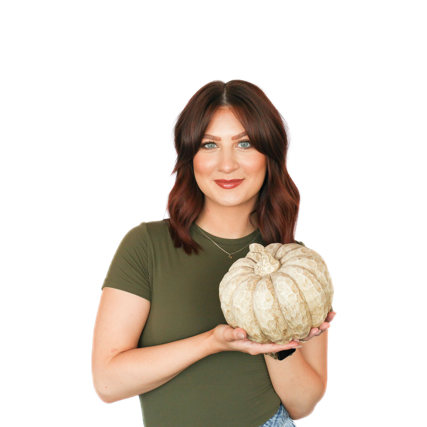 A woman with shoulder-length brown hair in a green shirt holds a pale, textured pumpkin with both hands against a white background.