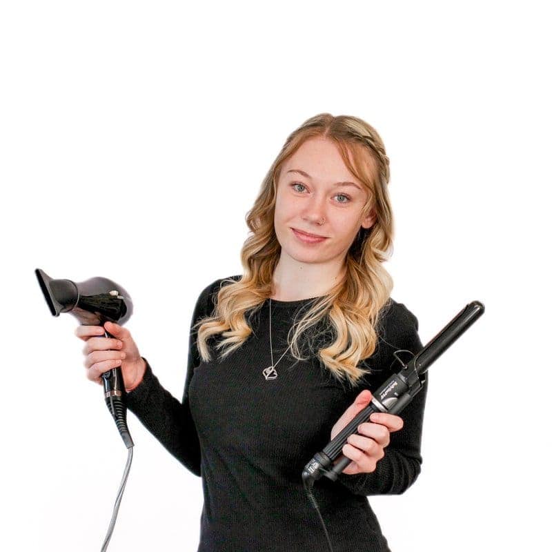 Woman holding a hairdryer in one hand and a curling iron in the other, smiling against a white background at a cosmetology school.