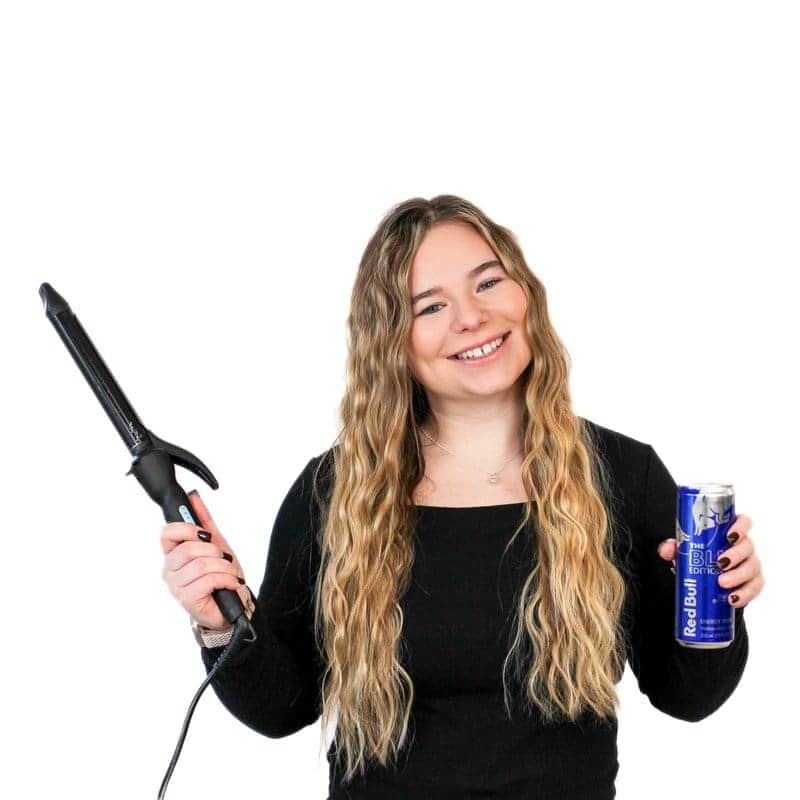 A young woman smiling while holding a hair curling iron in one hand and a can of hairspray in the other at a hair salon, against a white background.