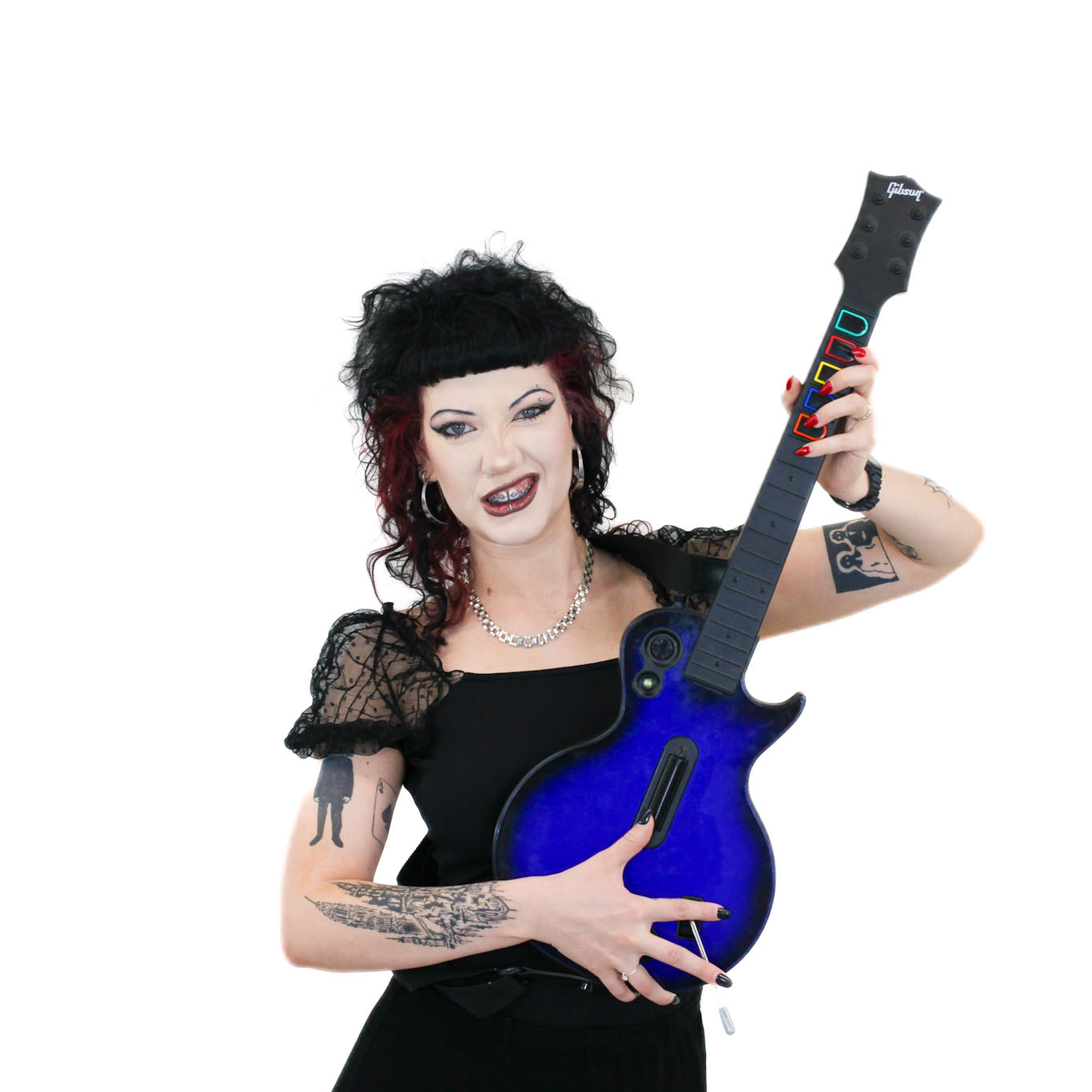Person with curly dark hair and tattoos holds a blue video game guitar controller, smiling and making a playful face against a white background.