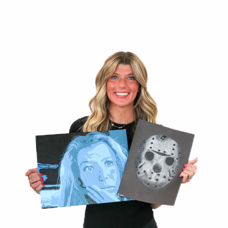 A person stands holding two artworks, one featuring a blue-toned portrait and the other showing a mask with multiple holes.