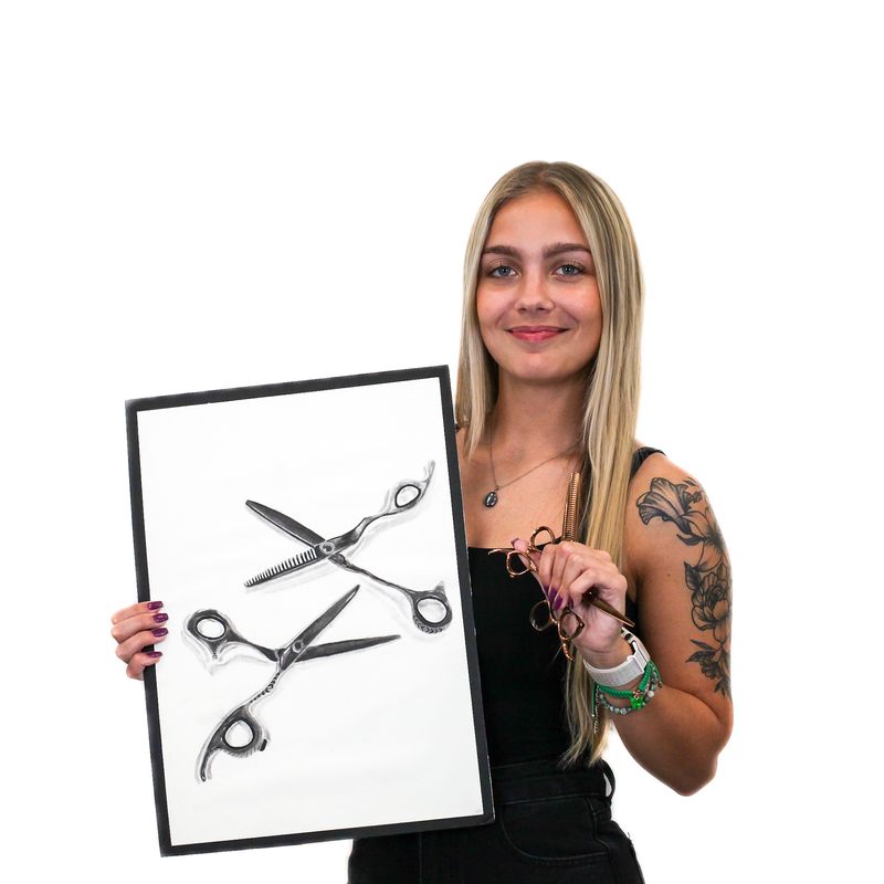 Woman with long blonde hair holding framed image of scissors in one hand and glasses in the other, smiling, wearing a black outfit, and showing tattoos on her right arm.