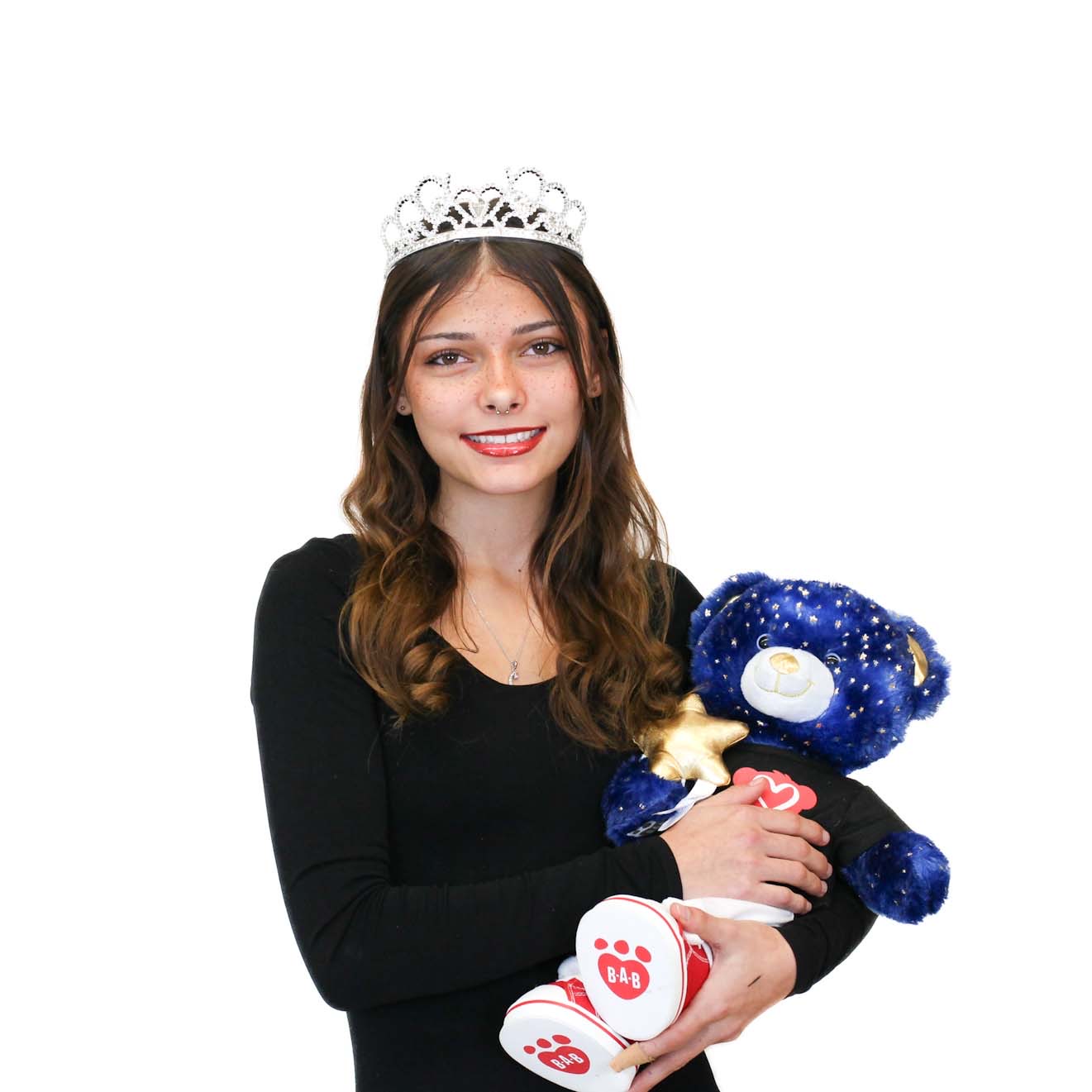 Young woman wearing a tiara and black top, holding a blue teddy bear with star patterns and a red heart on its chest, against a plain white background.