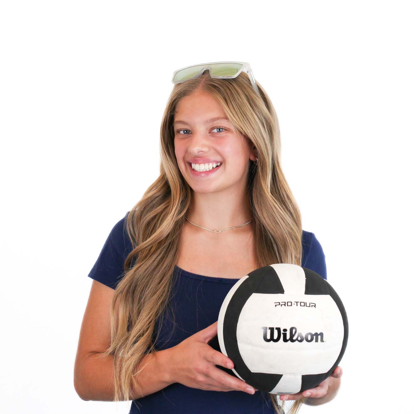 A young woman with long hair, wearing a navy shirt and sunglasses on her head, smiles while holding a black and white Wilson volleyball.