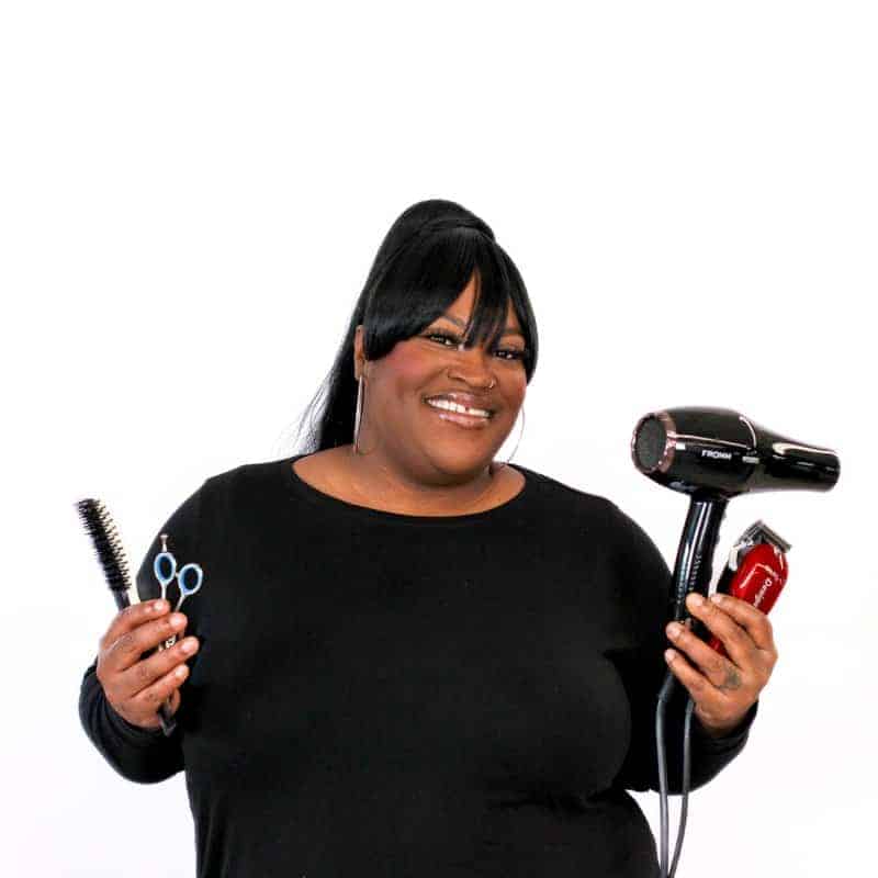 A smiling hairstylist at a hair salon in Omaha, holding a hairdryer, scissors, and a brush.