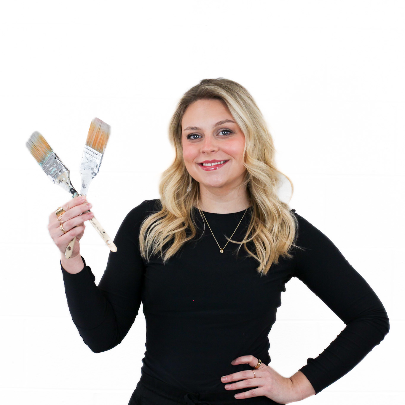 Woman with long blonde hair in a black top holds two paintbrushes in one hand and smiles, standing against a plain white background.
