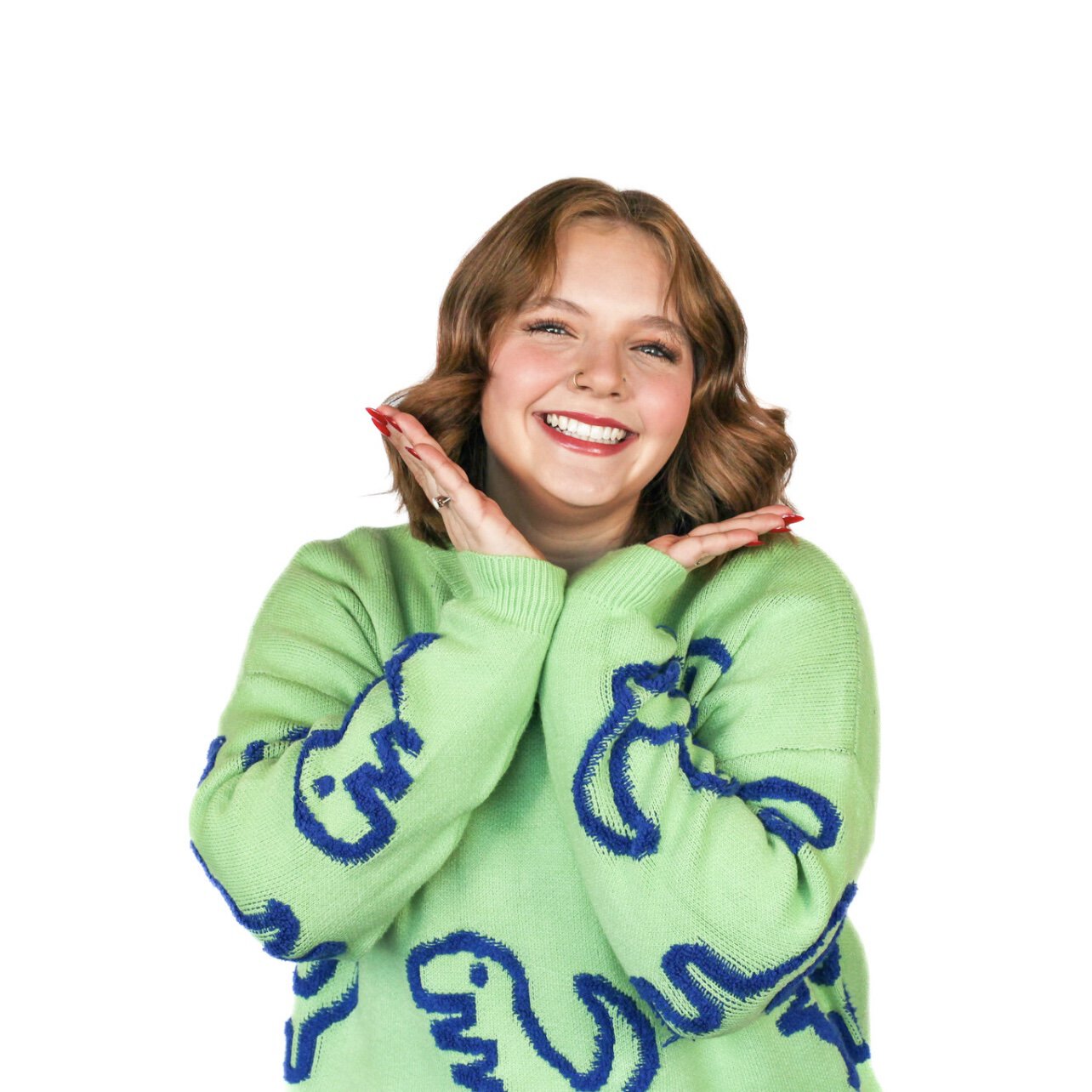 A smiling person wearing a green sweater with a blue dinosaur pattern, resting their face on their hands.