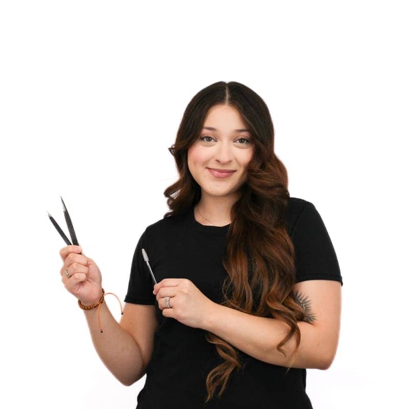 A woman with long wavy hair, trained at an Omaha hair school, holding scissors, smiling at the camera against a white background.