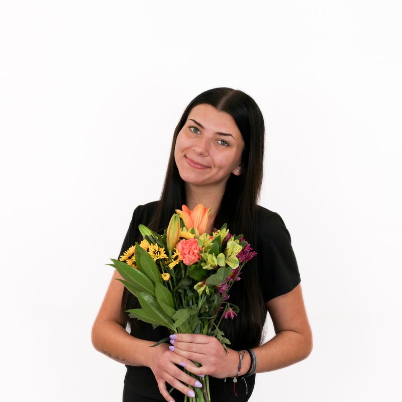 A woman with long dark hair holds a colorful bouquet of flowers, including orange lilies, pink carnations, yellow blooms, and green leaves, against a plain white background.