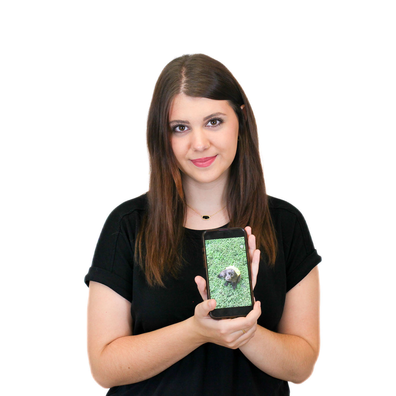 A woman with long brown hair in a black shirt holds a smartphone displaying a photo of a small dog on grass.