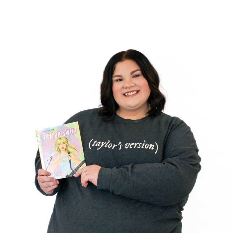 A person with dark hair and a gray "Taylor's version" sweatshirt is smiling and holding a "Taylor Swift" book with an illustrated cover, standing against a white background.