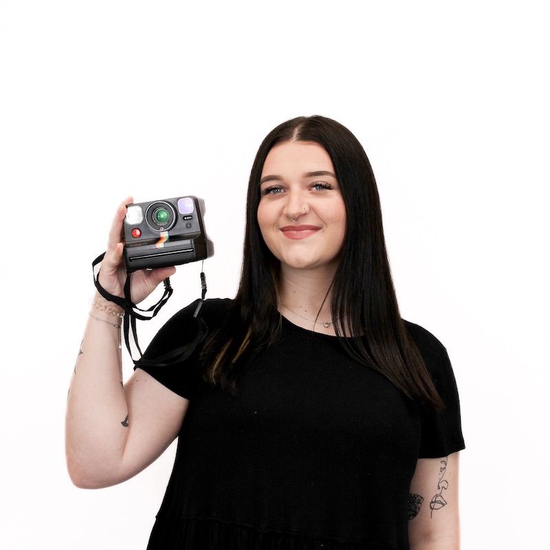 A person with long dark hair in a black shirt holds a Polaroid camera against a plain white background.