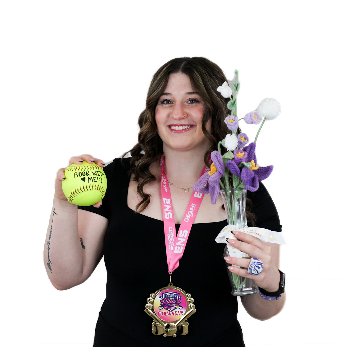 A woman in a black dress holds a yellow softball and a bouquet of knit flowers, wearing a pink lanyard, medal, and a championship ring against a white background.