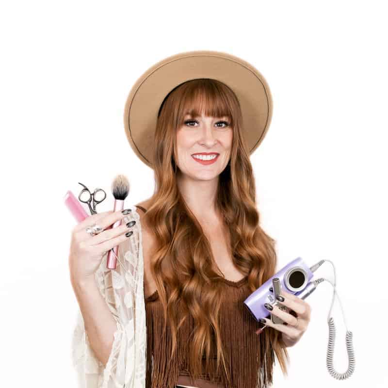 Woman smiling while holding makeup brushes and a compact camera, wearing a tan hat and a fringe top, fresh from the hair salon.