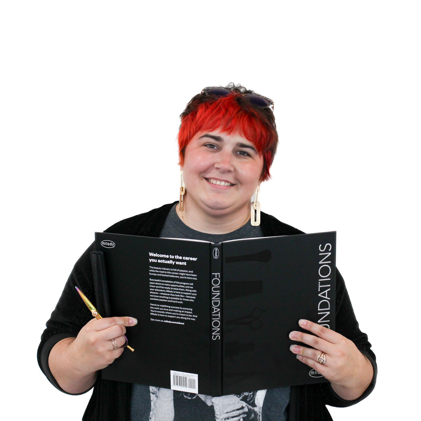 Person with short red hair and sunglasses on head smiling while holding an open book titled "Foundations" and a pen against a plain white background.