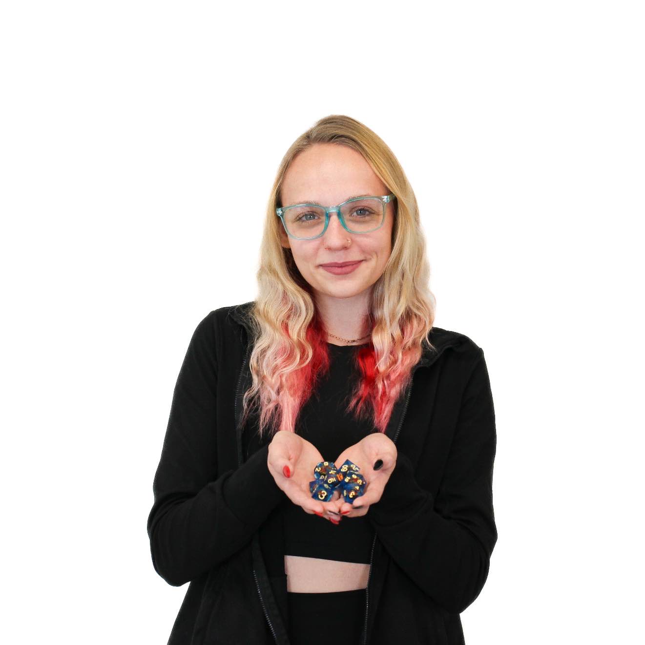A woman with blonde and red hair, wearing glasses and a black jacket, holds a collection of colorful polyhedral dice in her hands against a white background.