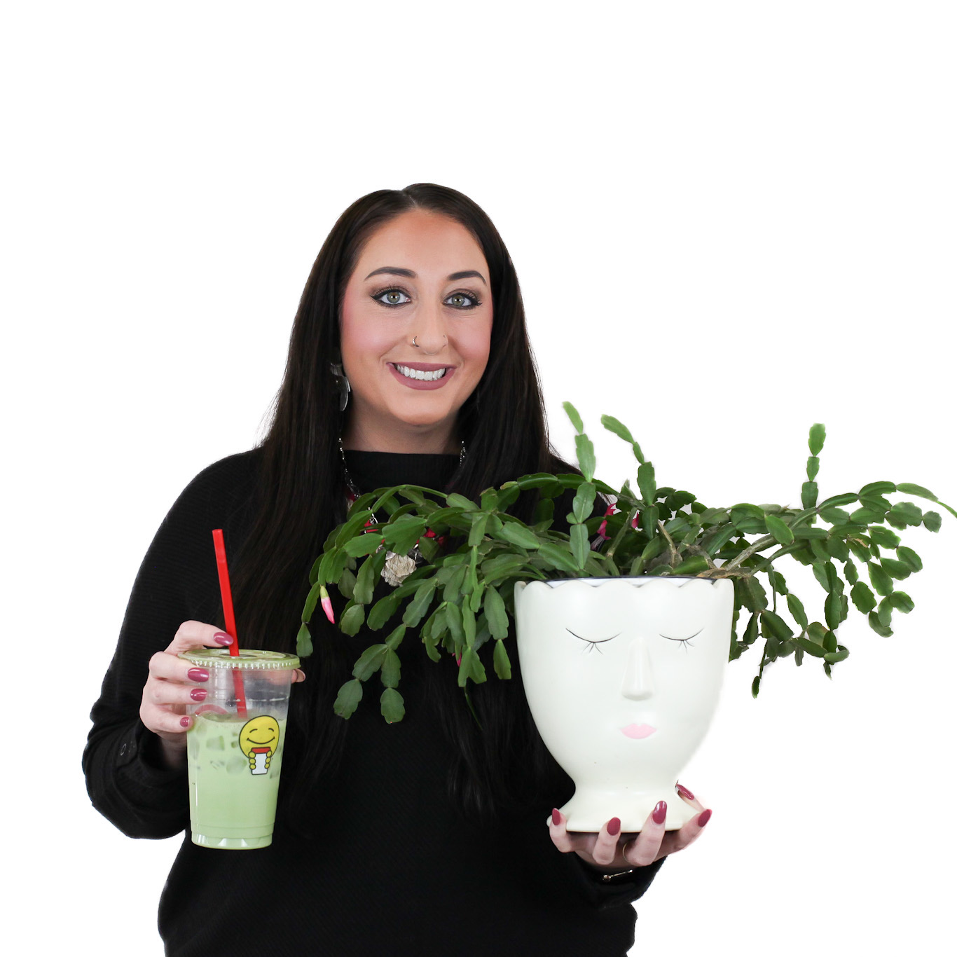 A woman with long dark hair holds a large potted plant shaped like a face in one hand and a green drink with a red straw in the other, standing against a white background.