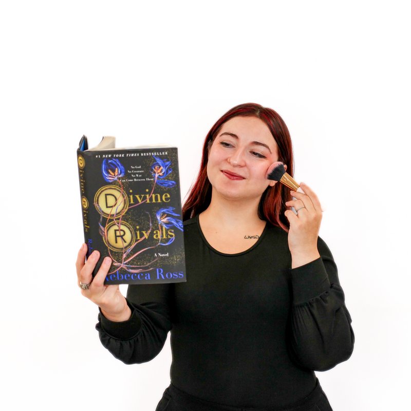 Person with medium-length red hair is applying makeup with a brush while holding and looking at a book titled "Divine Rivals" by Rebecca Ross. They are wearing a black top against a plain white background.