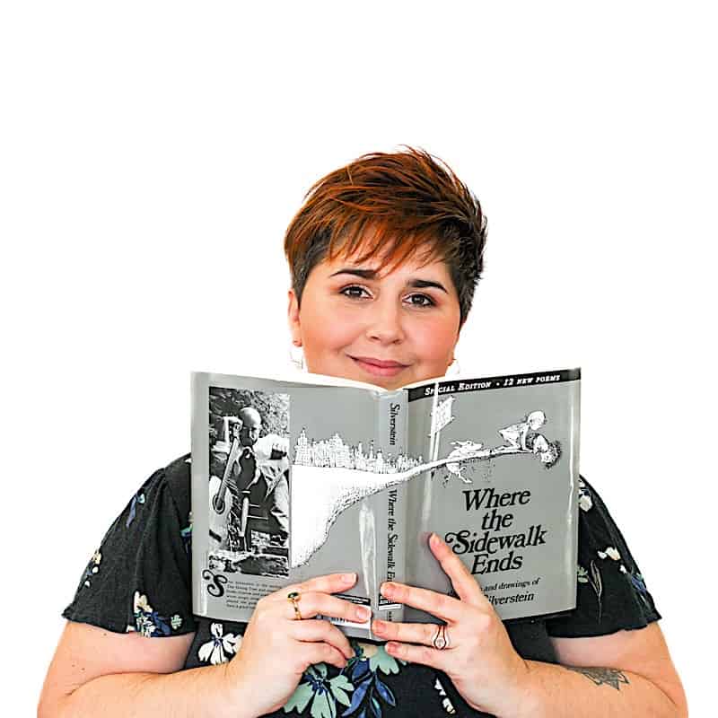 A person with short hair holds up and reads the book "Where the Sidewalk Ends" by Shel Silverstein, facing the camera, against a plain white background.